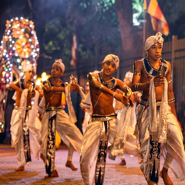 Group of male traditional Kandyan dancers in elaborate costumes and ves headdresses, playing geta beraya drums and holding torches, performing during the night procession of the Kandy Esala Perahera.