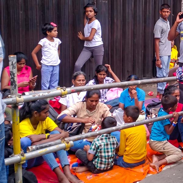 A crowd of spectators sitting on pavement mats behind street barricades, waiting to view the Kandy Esala Perahera