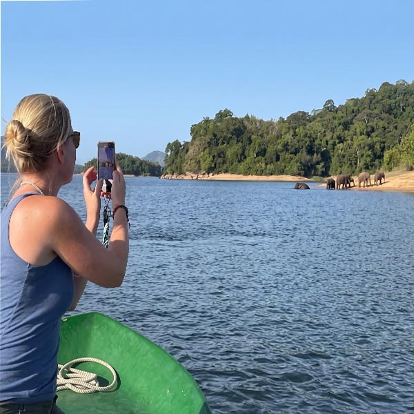 A tourist taking a photo from a boat during a scenic boat safari on the Senanayake Samudraya reservoir, with wild elephants visible on the distant shore in Gal Oya National Park.
