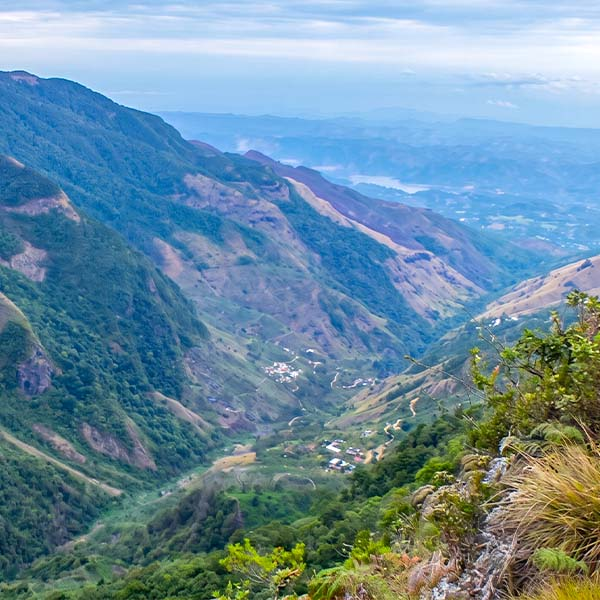 A dramatic view from a steep cliff edge, looking down a lush green mountainside into a deep valley with a small village below.