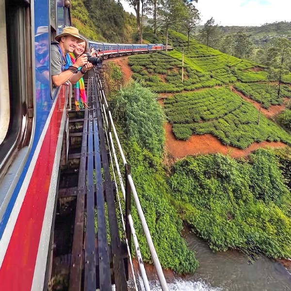 Smiling travelers leaning out the open doors of the iconic blue hill country train, passing through lush green tea plantations on their scenic journey from Ambewela to Ella.