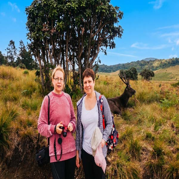 Two women posing for a photo near a nature museum before their hike, with a large Sambar deer standing in the grassy field behind them.