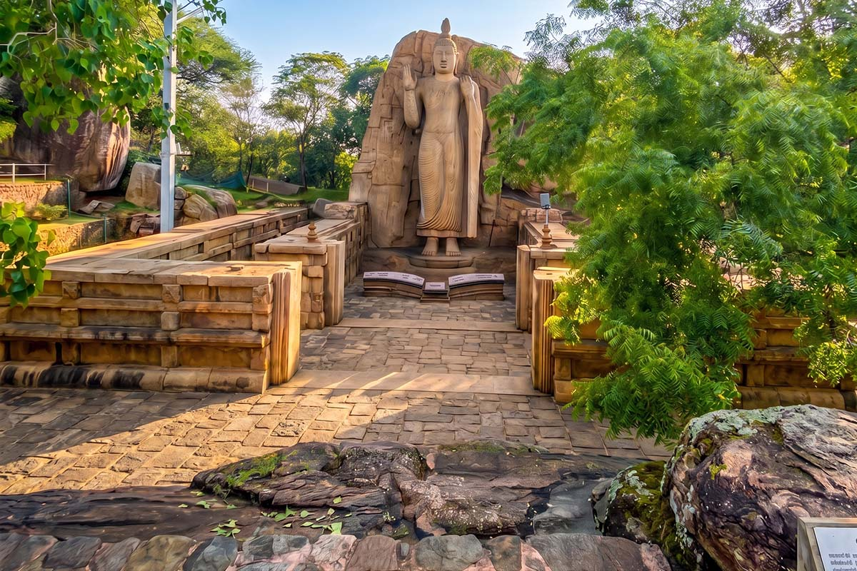 46-foot monolithic Awukana Buddha statue carved into a granite cliff face, surrounded by stone ruins and lush greenery under a clear sky.