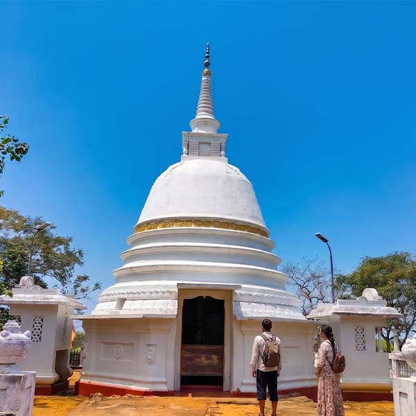 Visitors walking past a white bell-shaped stupa with a gilded spire under a blue sky at Awukana Temple.