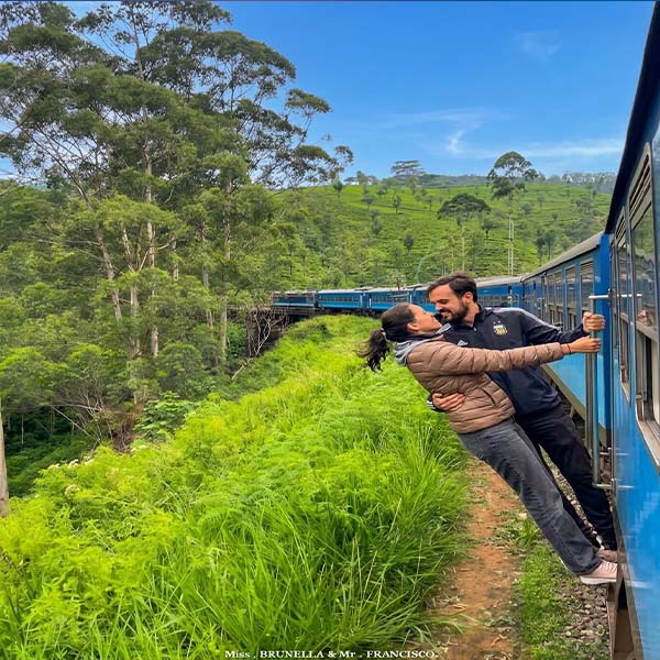 A couple leans out from an open doorway of a blue train as it travels through lush green, tea-covered hills in Sri Lanka, holding onto the handrails while leaning toward each other.