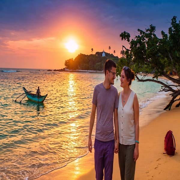 A couple stands together on a beach in Sri Lanka, looking out at the ocean at sunset. The setting sun casts a warm, orange glow across the water and the sandy shore. A traditional wooden fishing boat is resting on the sand nearby.