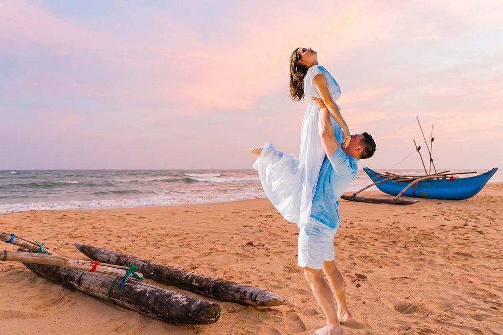 A groom lifts his smiling bride up into the air on a sandy Sri Lankan beach at sunset, with a traditional wooden outrigger boat resting on the shore nearby.
