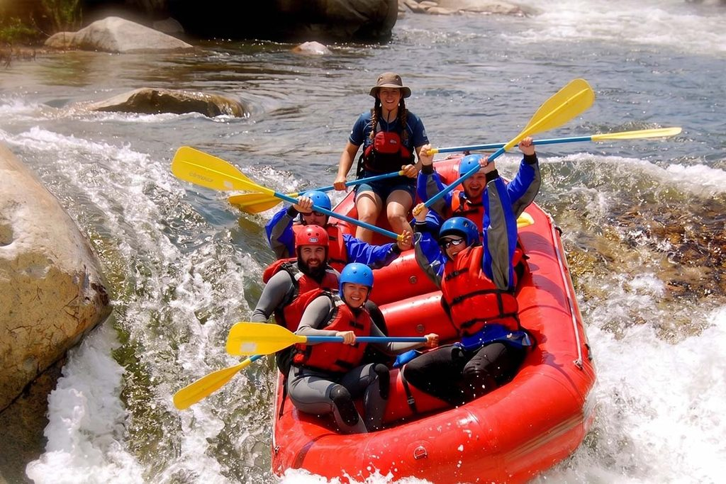 A group of people in a bright red raft navigating white water rapids on the Kelani River in Kitulgala, Sri Lanka, with one person holding a paddle up in the air.