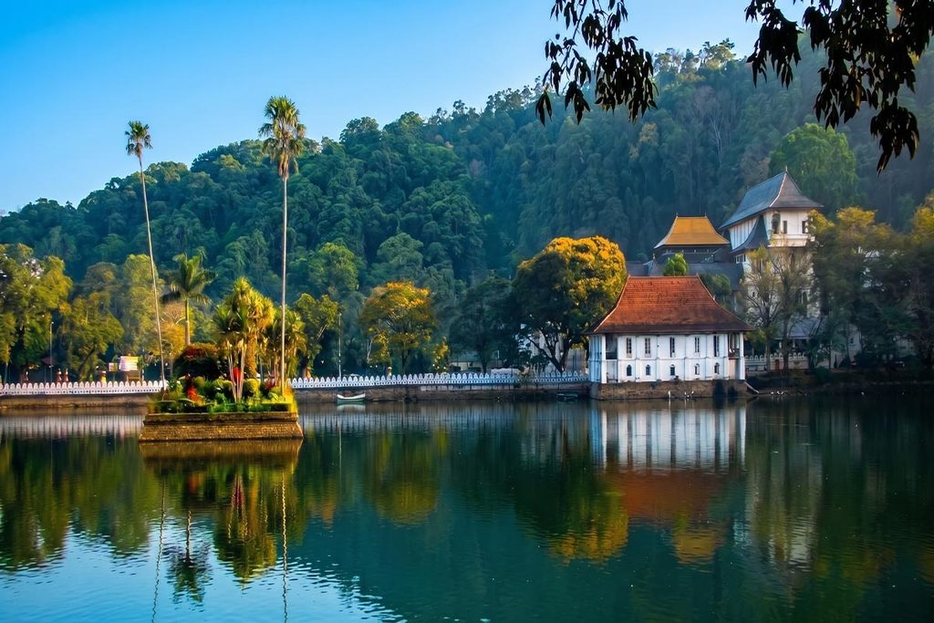 A scenic view of Kandy Lake reflecting the historic Temple of the Sacred Tooth Relic (Sri Dalada Maligawa) surrounded by lush tropical trees and traditional Sri Lankan architecture under a clear sky.