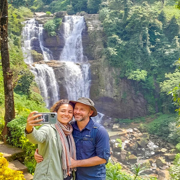 A scenic view of the rushing cascades of Ramboda Falls surrounded by lush green tropical forests, with two travelers smiling in the foreground.