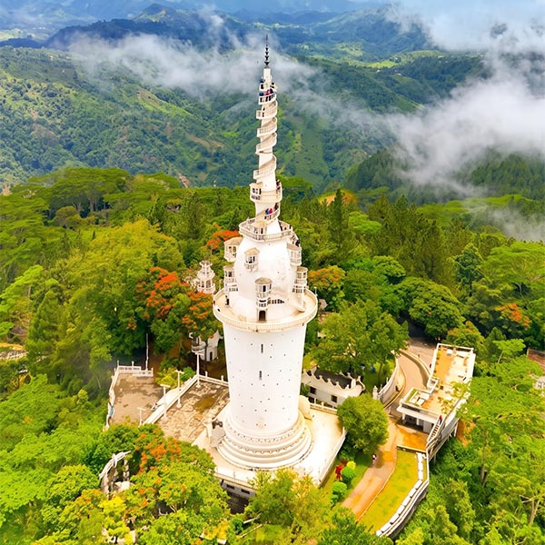 A scenic view of the Ambuluwawa Tower rising above misty green hills, with a traditional Sri Lankan building and lush tropical trees in the foreground under a cloudy sky.