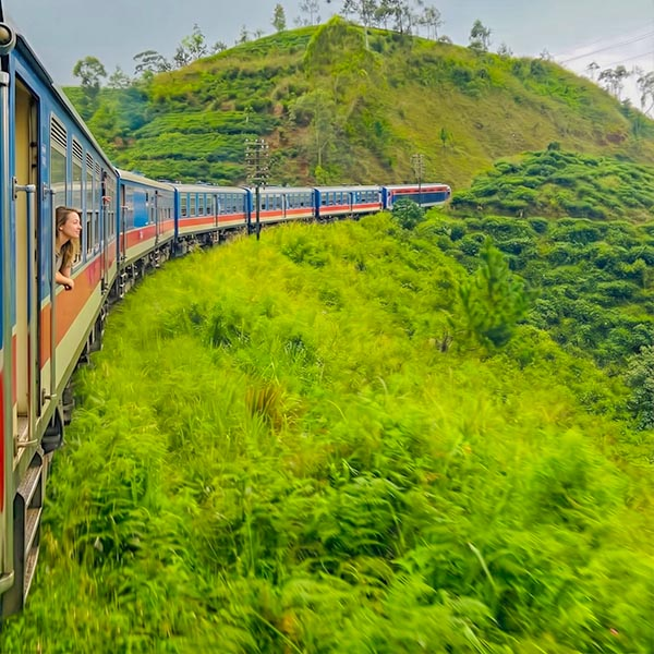 Travelers leaning out of a blue train window as it winds through lush green hills along the Sri Lankan highlands railway line