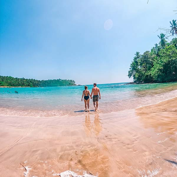 Couple holding hands walking along the sandy shore of Hiriketiya Beach with turquoise ocean waves and lush green trees in the background.