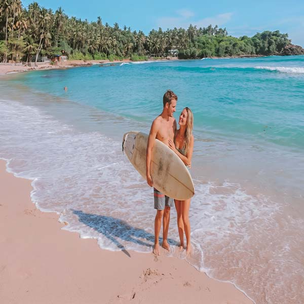 Couple holding a surfboard on the sandy beach of Hiriketiya  with clear turquoise ocean waves in the background.