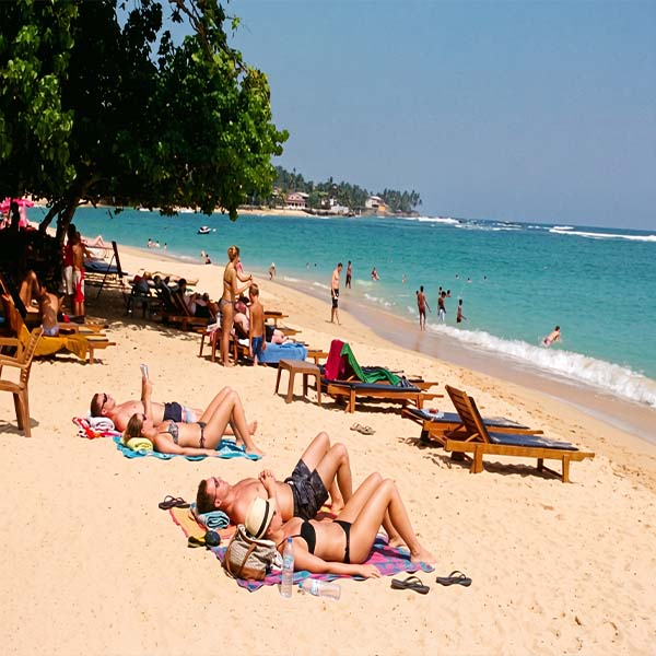 Sunbathers relaxing on the golden sand of Unawatuna Beach with turquoise ocean waves and sun loungers under palm trees.