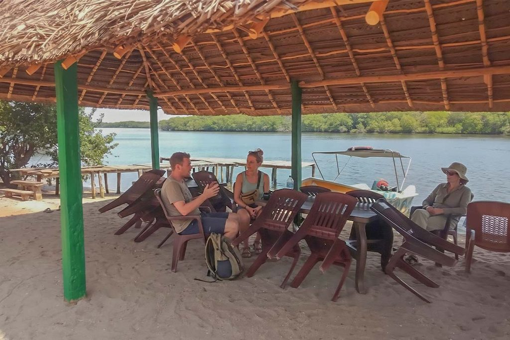 Travelers relaxing under a thatched-roof shelter with a view of the Kala Oya river and mangrove trees during a boat cruise in Gange Wadiya, Sri Lanka.