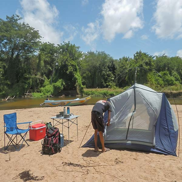 Travelers camping overnight in a tent on the sandy banks of the Kala Oya river next to a blue folding table and chairs in Gange Wadiya, Sri Lanka.