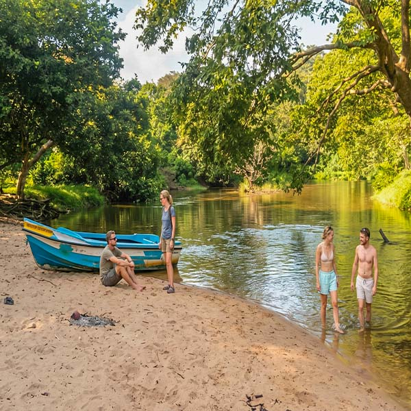 ravelers relaxing on a boat along the Kala Oya riverbank with a blue canoe, lush green mangrove forests, and a scenic water channel in Gange Wadiya, Sri Lanka.