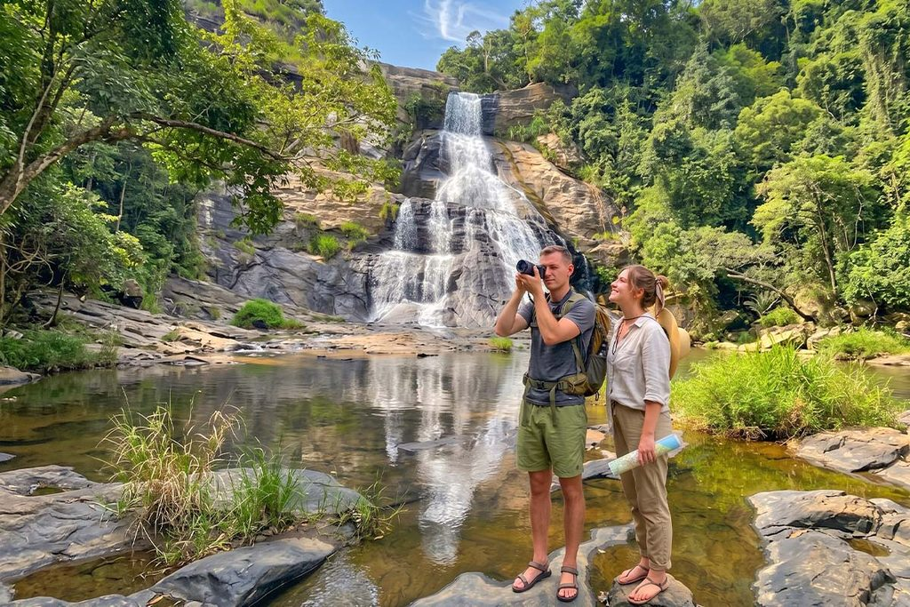 Two travelers taking photographs of a scenic waterfall and mountain stream during a guided day trip in Kandy.