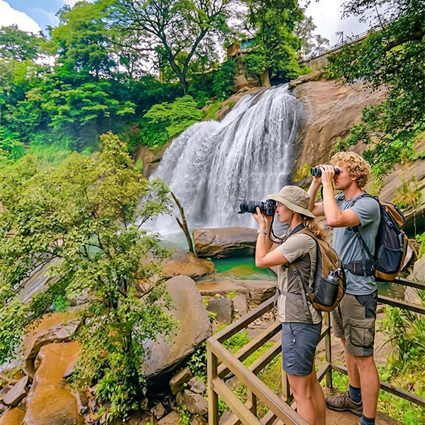 Two travelers taking photographs of a scenic waterfall and mountain stream during a guided day trip in Kandy.